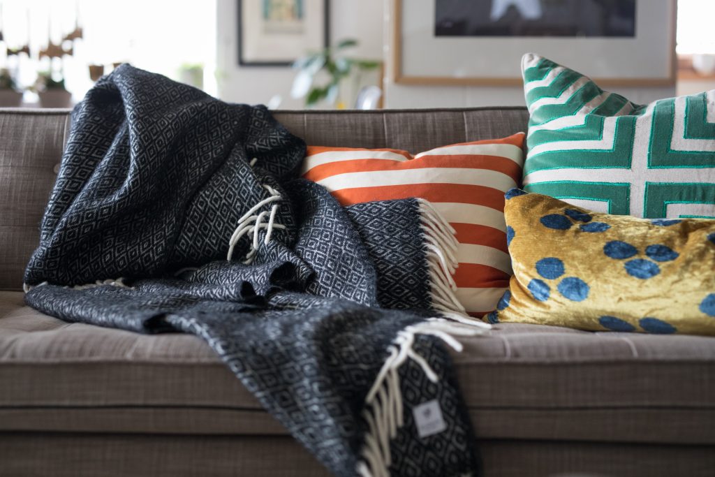 Ein graues Sofa mit einer dunkelgrauen Kuscheldecke und 3 bunten gemusterten Kissen.