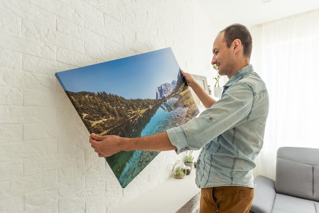 Die Verjüngungskur für das eigene Zuhause: 7 coole Ideen 1 Leinwanddruck mit Galeriefolie und Innendekor aus trockenem Gras. Frau hängt Fotografie an weiße Wand. Händehaltender Fotoleinwanddruck mit Bild einer Insel in der Stadt Biarritz in Frankreich. Foto in hoher Qualität