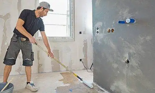 Ein Mann grundiert mit einer Grundierrolle an einer Teleskopstange den Boden im Badezimmer.