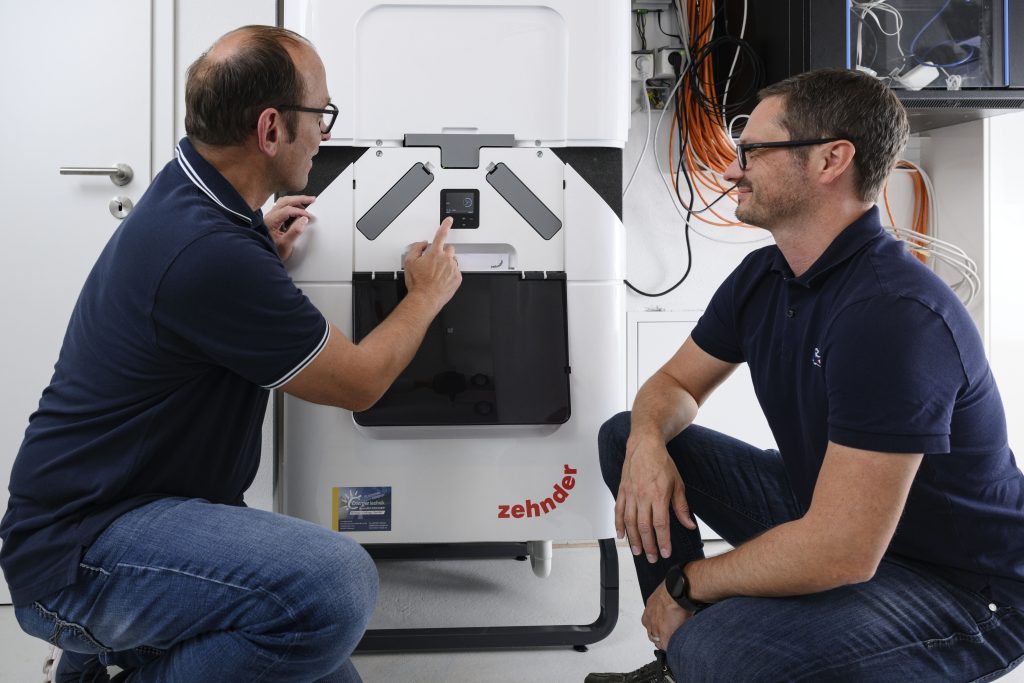 Zehnder ComfoClime Cool. Ein Mann zeigt einem anderen Mann wie die Kühleinheit Zehnder Comfo Clime Cool von Zehnder funktoniert. Beide knien vor dem grau-weißen Gerät. Die Männer tragen dunkelblaue Polohemden. Sie befinden sich im in einem Keller- oder Technikraum.