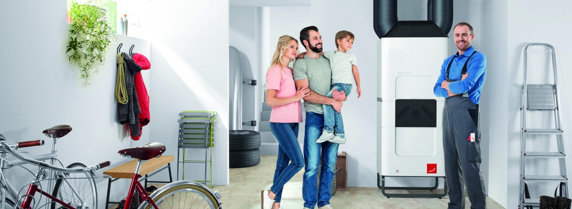 Kühlung Einfamilienhaus. Eine Familie steht mit Monteur lächelnd im Heizungskeller. Dort werden auch Fahrräder aufbewahrt. Der Vater hat das Kleinkind auf dem Arm.