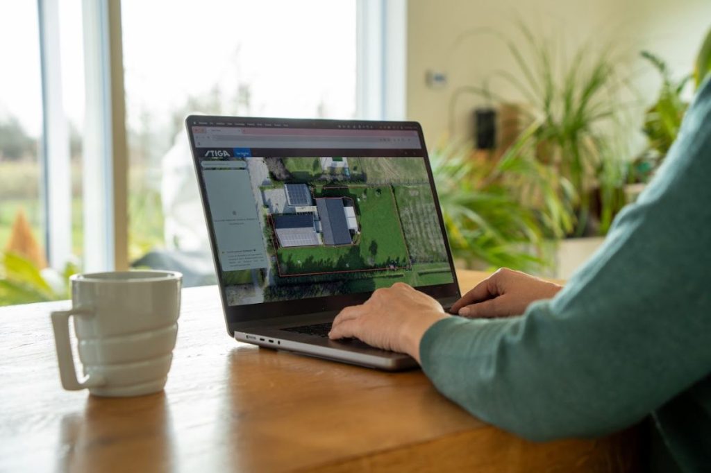 Stiga Rasenroboter einstellen am PC. Ansicht einer Person, die am Laptop sitzt. Daneben eine Tasse auf dem Tisch. Im Hintergrund unscharf ein Terrassenfenster und Zimmerpflanzen.