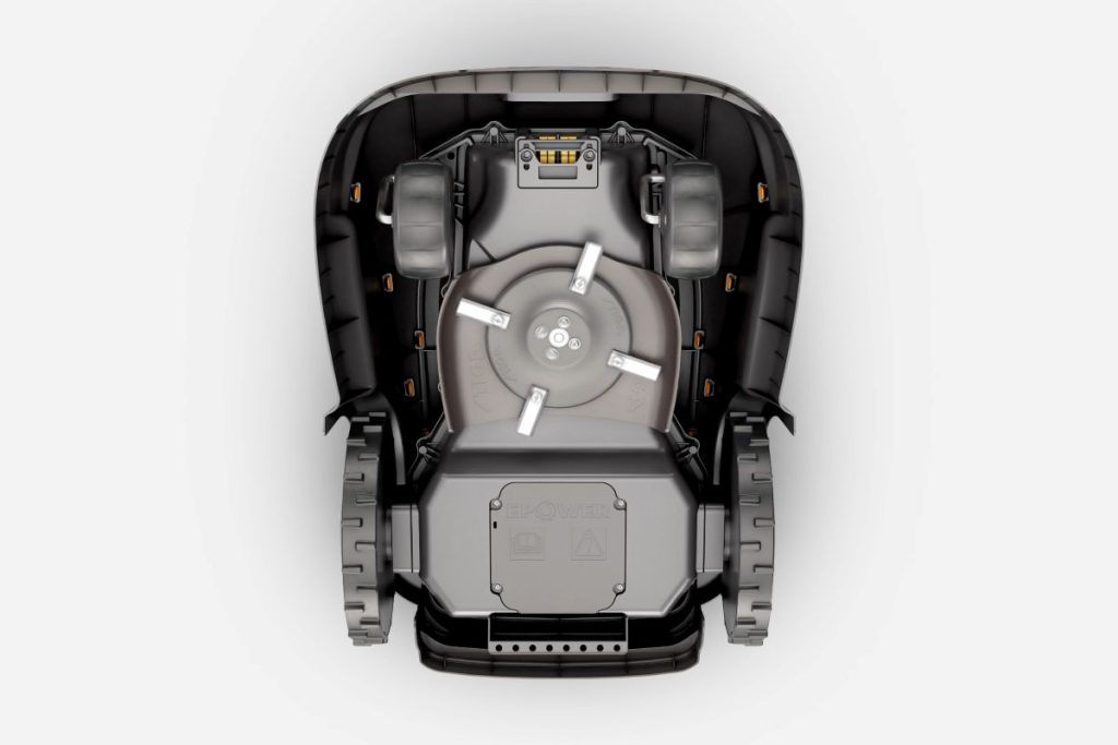 Stiga Rasenroboter von unten: Stiga Rasenroboter dunkel: Detailansicht des Rasenroboters. Der Roboter ist anthrazit und hat einen roten Schalter. Orangefarbig abgesetzte Elemente am Gehäuse. Deutlich zu sehen die zwei kleineren Vorderräder und die großen Hinterräder.