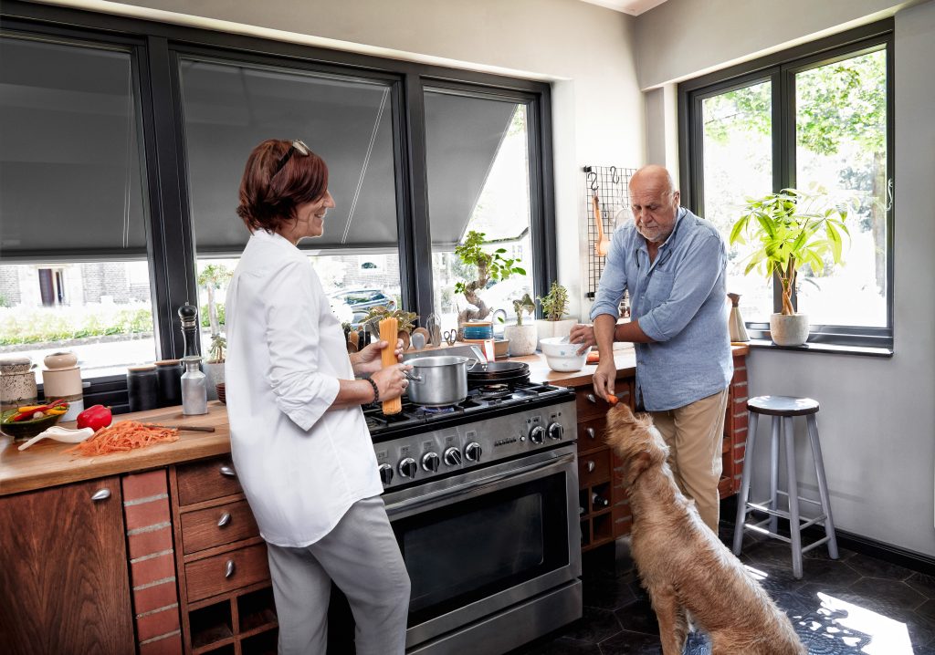 Ein älteres Ehepaar in der Küche bei der Zubereitung des Abendessens. Der Mann füttert einen Hund. An den Fenstern ist ein herunter gelassener Sonnenschutz zu sehen. Warema Sonnenschutz Markise