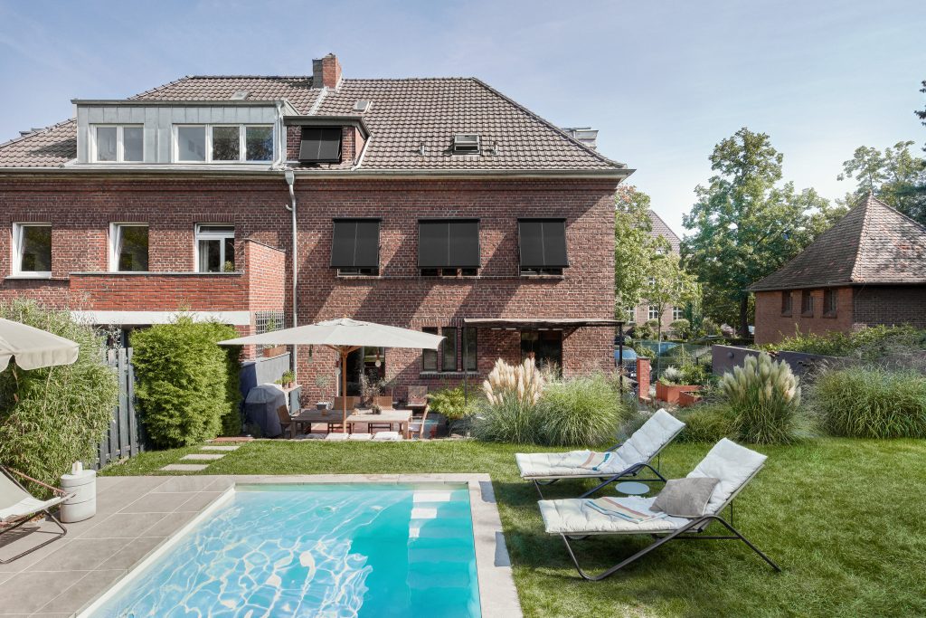Warema Sonnenschutz Markise. Ein Einfamilienhaus mit Sonnenschutz. Davor ein Garten mit Pool und Liegen dabei.