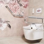 Renovieren – umfassender Ratgeber für Ihr Zuhause 3 Barrierefreie Toilettenanlage: Weißes Wand-WC mit integriertem Toilettenpapierhalter am seitlichen, hochklappbaren Stützgriff. An der angrenzenden Wand befindet sich ein winkelbarer Haltegriff vor einer Tapete mit großflächigen, altrosa Pfingstrosen-Illustrationen. Die Fliesen sind in hellem Beige gehalten.
