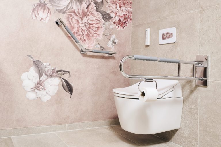 Themen 3 Barrierefreie Toilettenanlage: Weißes Wand-WC mit integriertem Toilettenpapierhalter am seitlichen, hochklappbaren Stützgriff. An der angrenzenden Wand befindet sich ein winkelbarer Haltegriff vor einer Tapete mit großflächigen, altrosa Pfingstrosen-Illustrationen. Die Fliesen sind in hellem Beige gehalten.