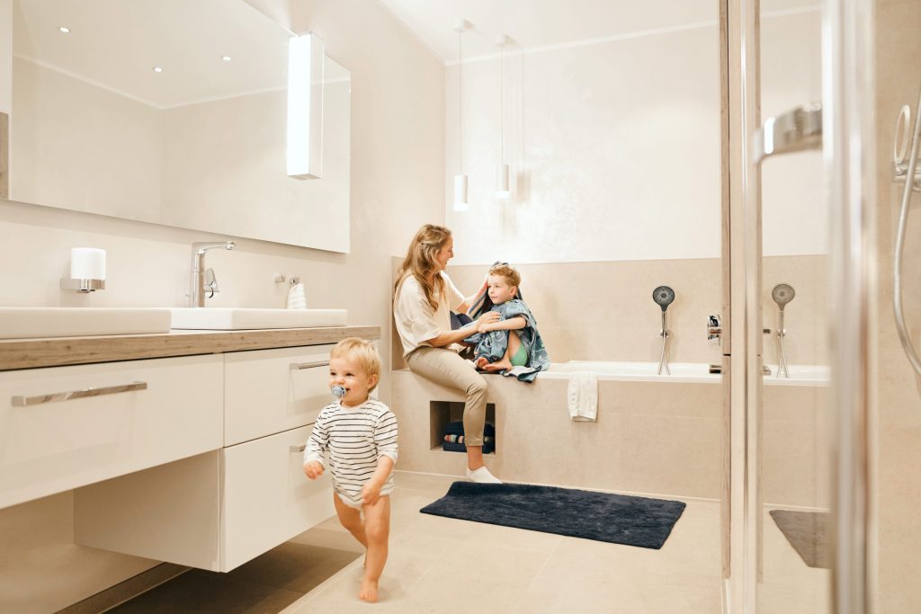 Traumbäder ohne Grenzen 6 Ein großzügiges, hell beige gefliestes Badezimmer. Eine Frau sitzt am Rand einer eingebauten Badewanne und trocknet ein Kind mit einem Handtuch ab, während ein Kleinkind im gestreiften Body im Vordergrund läuft. Links befindet sich ein langer weißer Waschtisch mit großem Spiegel und modernen Chrom-Armaturen. Die Beleuchtung ist sanft und wohnlich.